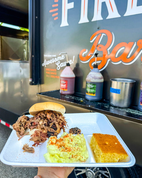 Tray of pulled pork sandwich, potato salad, and cornbread in front of a food truck.